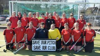 Southwick's women's team lined up in the goal mouth of their pitch with their hockey sticks and red tops with a sign that reads "save our pitch, save our club".