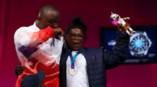 Person wearing glasses, a dark quilted jacket and a silver medal on a blue ribbon stands beside another person in a white and red sports jacket. The medal wearer raises a colourful mascot toy while standing on a sports event stage with a red backdrop.