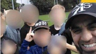 A man wearing a baseball cap takes a selfie with a group of seven children against a backdrop of sports pitches. The faces of all the children have been blurred.