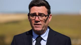 Mid shot of Andy Burnham wearing a black suit and black tie with black-framed glasses. In the distance is the white cliffs of Dover.