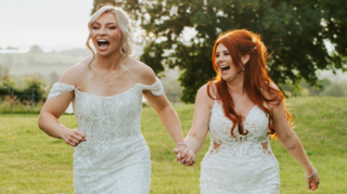 Lauren Price and Carlie Jones walking hand in hand in a field both laughing in white gowns