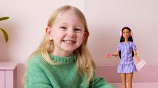 A young girl with blonde hair, smiling. She has a light green knitwear jumper on and is next to a Barbie doll which has ear defenders on and is holding a card and a fidget spinner.