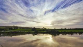 A blue sky filled with white clouds fanning out from the sun which is centred low over a field with a lake in front