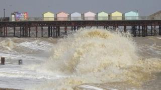 A row of pastel coloured beach huts sit on a pier as a large wave in the sea crashes in the forefront. The sky is grey in the backdrop.