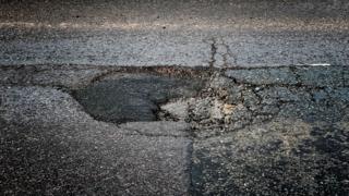 Close-up image of a pothole. It is roughly circular with a number of cracks, and part of it looks to have been filled with asphalt, which is darker in colour than the surrounding road surface.