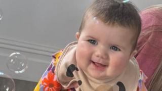 A smiling female baby looking at bubbles being blown in her direction. She has brown hair and blue eyes. She's wearing a beige bib and a colourful, floral top.