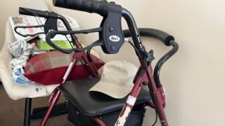 A zimmerframe is placed in the corner of a care home. The zimmerframe has a maroon colour and has a beige cap on the seat. There is a seat next to it with a newspaper.