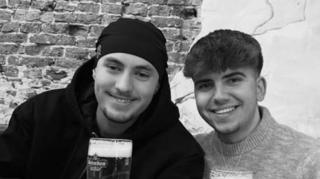 A black and white photo of Eddie and Jayden, smiling side by side. They are both holding pints or beer. Jayden has a hat on and has short stubble and is wearing a black hoodie. Eddie has short dark hair and is wearing a jumper.