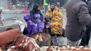 Two earthquake survivors sit wrapped in blankets in front of a makeshift fire