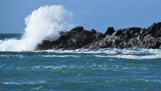 A wave crashes over rocks with a bright blue sky in the background in Isle of Colonsay, Scotland