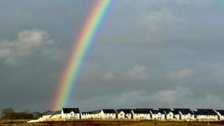 A large vibrant rainbow rises from left over a row of white houses with black roofs. The sky is grey and cloudy