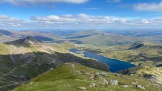 A thin layer of white clouds float over a Welsh mountain range and lake, casting their shadows on the ground as they pass.