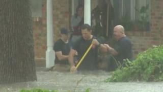 Three rescuers, waist-deep in flood water, help residents from their home.