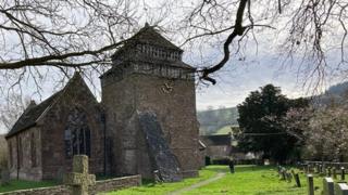 A church and a graveyard with bare tree branches in front and lightly cloudy skies behind.