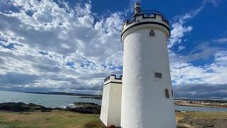 A lighthouse painted bright white stands out against blue skies with fluffy clouds , with the sea behind.