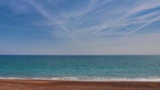 A straight stretch of beach with the green/blue sea behind and light white clouds in a blue sky