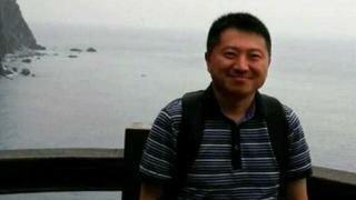 Liu Hu wearing a blue polo shirt and smiling as he poses for a photo. Behind him is the sea.