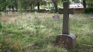 A gravestone in a cemetery, with other gravestones to the left. There is a lot of overgrown grass around the graves and trees and a brick wall in the distance. A building is also behind the graves.