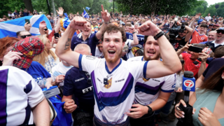 Crowd of sports fans in a park waving Scottish flags, with a central person in a white and purple jersey raising both fists while surrounded by reporters and onlookers.
