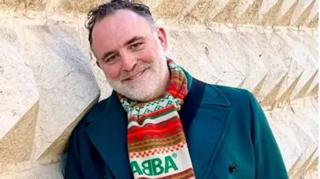 The image features a man wearing a dark green coat and a vibrant scarf. The scarf is patterned with red, green, and white colours, prominently displaying the text "ABBA" on it. Behind them is a textured stone wall with intricate geometric designs