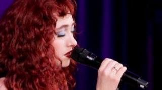 Singer Chappell Roan looking sombre as she sings into a microphone. She has red hair and pale complex. There is a ring on her wedding finger. The background is dark purple. 