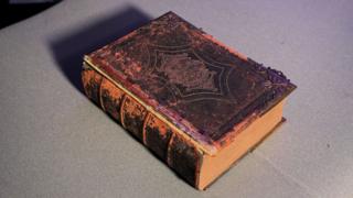 Ar ford llwyd mae hen Feibl teuluol, gyda marciau amlwg ar y clawr lledr yn dangos oedran sylweddol y gyfrol. Mae lliw oren a phorffor ar ymylon y clawr, gyda'r lledr brown dal yn amlwg ar ganol y llun.