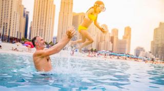 A man in the water at the beach in Dubai throwing his young daughter in the air. There are skyscrapers in the background