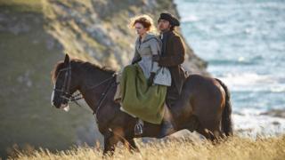 Ross and Demelza Poldark are sitting on a chestnut horse which is walking through long grasses on a sunny day. Behind them is a sheer cliff and sparkling sea. Demelza is riding side saddle and is wearing a long dress, cloak and gloves while Ross is wearing a tricorn hat, heavy overcoat and gloves.