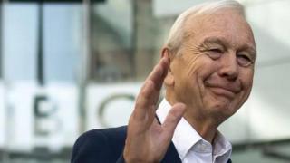 John Humphrys waving goodbye outside BBC's New Broadcasting House. He has white hair and is wearing a white shirt with a navy blazer.