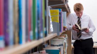 A GCSE student wearing uniform reads a book in a school library leaning against a shelf of books.