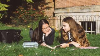 Two teenage schoolgirls in uniform lie on the grass reading and studying outdoors after class - stock image