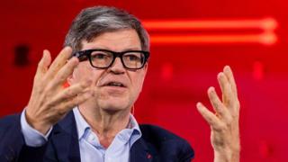 Yann LeCun wears fashionable thick-rimmed glasses, a light blue shirt and navy jacket. He is using his hands to articulate a point while speaking at a conference.