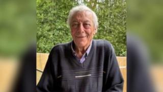 An elderly man with grey hair and wearing a dark blue jumper smiles at the camera