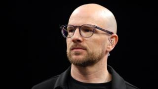 A close up of Mark Webster in dark shirt and jacket wearing spectacles and a TV earpiece