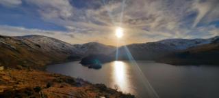 Sun shines in a partly cloudy sky above snow-capped mountains next to a lake