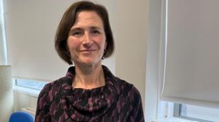 Annette Morris has short, brown hair wearing a dark purple and black patterned dress with a cowl neck in an office setting. 