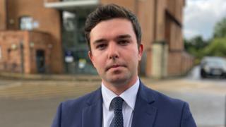 A dark-haired man wearing a navy suit and navy and white spotty tie and white shirt. He is standing in front of a blurred red-bricked building.