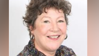 Sue Riley - a middle aged white woman with short, brown, curly hair. She is posing for a staged photo in front of an all-white background and is wearing a predominantly brown multi-coloured shirt. She has large hooped earrings on and is smiling