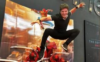A man jumps up and strikes a pose with his arms outstretched. He is in front of a sign reading 'Spider-Man - Brand New Day' against a grey cinema lobby wall