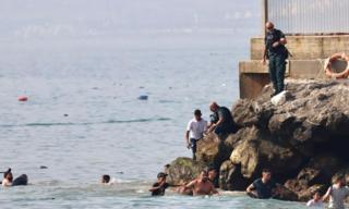 Men swimming as police officer standing on rocks holds arm of man in white t-shirt