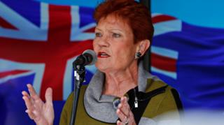 Pauline Hanson speaks into a microphone at a rally, an Australian flag in the background behind her