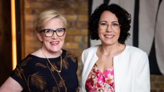 With blonde hair and wearing a black blouse Monika Liikamaa stands next to Denise Johansson who wears a white jacket and flowery pink blouse.