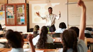 A male teacher stands at the front of a classroom facing towards the students. Some students are raising their arms in the air.