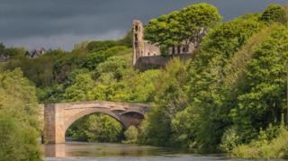 The ancient, ruined castle and its bridge across the River Tees. It is lit by warm, afternoon sunlight - but the sky is a deep, dark grey. Bright green trees with huge canopies of leaves partially obscure the castle and spread out from both banks of the river.