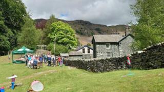 Langdale CofE Primary School in the Lake District, Cumbria. The school building is built with picturesque blue slate. It is in a green environment and has a dry stone wall surrounding the premises. Its grassy grounds are filled with parents and children on what looks like a sports day.