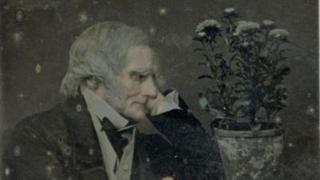 The image shows an elderly man with grey hair wearing a suit and looking at a pot of flowers.