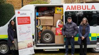 An ambulance with its side door is open. Lots of cardboard boxes and plastic bags are stacked on the inside and a spare tyre can be seen. Three people, a man and two women stand next to it.