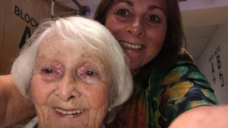 Two woman take a selfie. The woman on the left has short white hair and green eyes. The woman on the right, who takes the picture, has short brown hair and brown eyes, she is wearing a green and yellow patterned top.