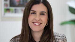 A woman with long dark hair and hoop earrings, wearing a check blazer, smiles at the camera.