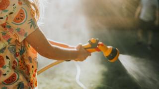 A girl wearing a patterned top holds a yellow garden hose with an attachment. She sprays water from the hose attachment. A man stands in the top right background of the picture.